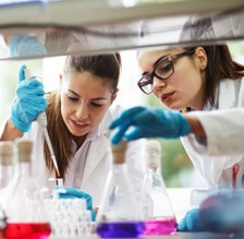 Two women in a lab, dressed in lab coats and wearing blue gloves.