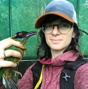 a woman outdoors, wearing a hat and glasses, and holding a bird